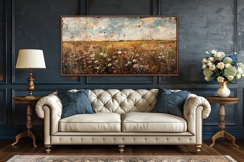 Puede incluir: Una pintura al &oacute;leo de un campo de flores silvestres en un marco marr&oacute;n cuelga sobre un sof&aacute; crema con dos cojines azules. Una l&aacute;mpara dorada y un jarr&oacute;n de flores se encuentran a ambos lados del sof&aacute;. La pared es azul oscuro.