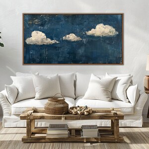 May include: A white couch with white pillows in front of a blue and white abstract painting with three white clouds. The couch is in a living room with a wooden coffee table and a wooden side table with a lamp.