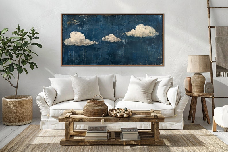May include: A white couch with white pillows in front of a blue and white abstract painting with three white clouds. The couch is in a living room with a wooden coffee table and a wooden side table with a lamp.