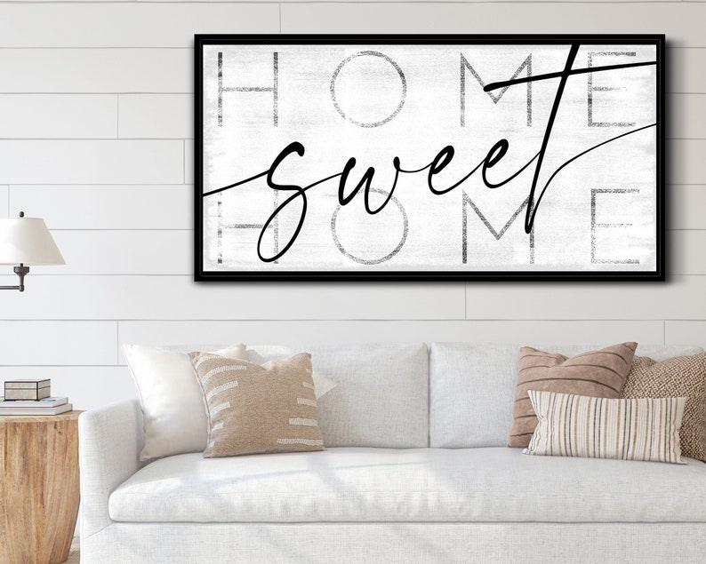 May include: A framed wall art piece with the words "HOME sweet HOME" in black script and block letters. The background is a distressed white, and the frame is black. The artwork is displayed above a white sofa with decorative pillows.