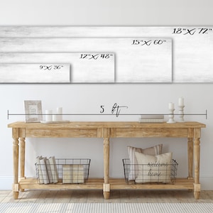 May include: A whitewashed wood wall with four different sized rectangular canvases hanging on it. The canvases are labelled with their dimensions in inches. The wall is above a wooden console table with two black wire baskets on the bottom shelf. The table is 5 feet long.