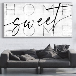 May include: A rectangular canvas art piece with the words "HOME sweet HOME" in black script and block letters against a white background. The artwork hangs above a gray sofa with pillows.
