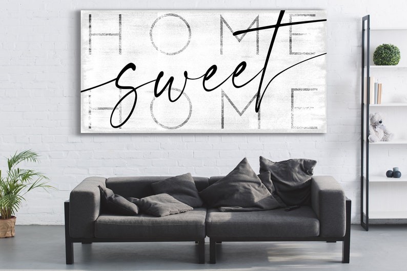May include: A rectangular canvas art piece with the words "HOME sweet HOME" in black script and block letters against a white background. The artwork hangs above a gray sofa with pillows.