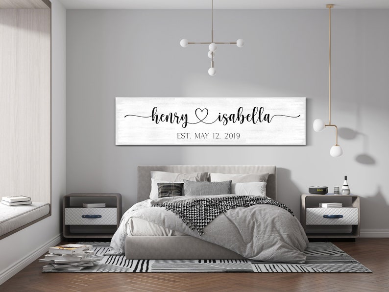 May include: A white wooden sign with black calligraphy that reads "henry &hearts; isabella est. may 12, 2019" hanging above a grey upholstered bed with white bedding and a black and white throw blanket.
