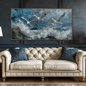 Puede incluir: Un sof&aacute; de cuero blanco con botones capitonn&eacute;s y dos cojines azules se encuentra frente a una pared azul oscuro con una pintura enmarcada de gaviotas volando sobre un mar tormentoso. La pintura est&aacute; en tonos de azul, blanco y gris.