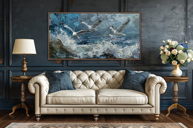Puede incluir: Un sof&aacute; de cuero blanco con botones capitonn&eacute;s y dos cojines azules se encuentra frente a una pared azul oscuro con una pintura enmarcada de gaviotas volando sobre un mar tormentoso. La pintura est&aacute; en tonos de azul, blanco y gris.