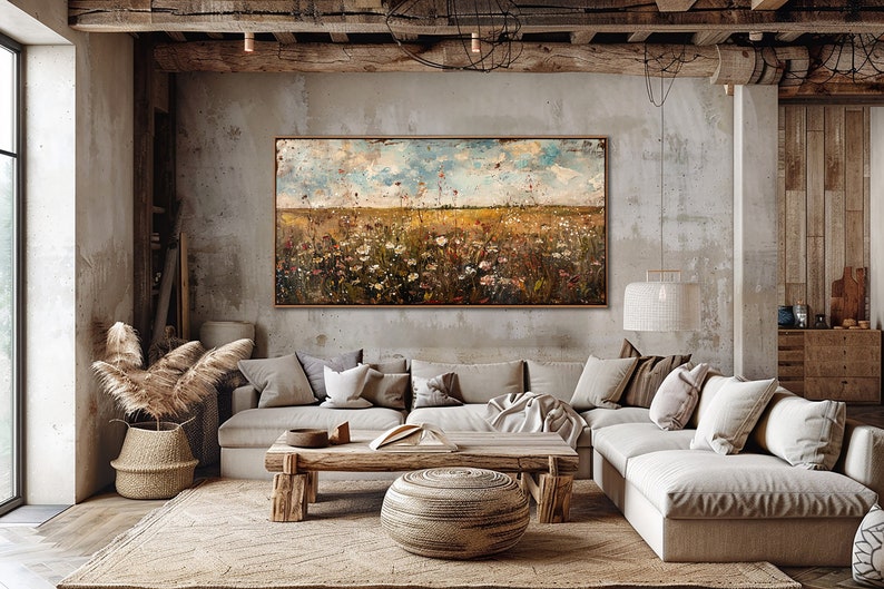 Puede incluir: Pintura de paisaje abstracto con un campo de flores silvestres bajo un cielo azul y nublado. La obra de arte est&aacute; enmarcada en madera oscura y cuelga sobre un sof&aacute; seccional beige. La habitaci&oacute;n tiene una est&eacute;tica r&uacute;stica y natural.