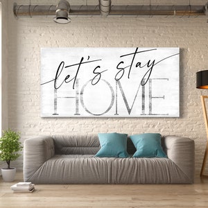May include: A white canvas wall art print with black text that reads "Let's stay home". The print is hanging on a white brick wall above a gray couch with blue throw pillows. A wooden floor lamp with a black lampshade is in the corner of the room.