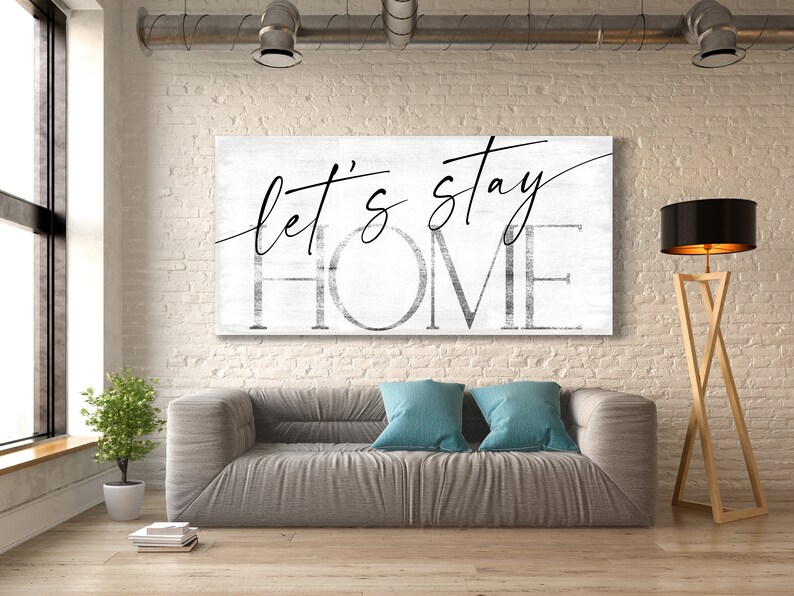 May include: A white canvas wall art print with black text that reads "Let's stay home". The print is hanging on a white brick wall above a gray couch with blue throw pillows. A wooden floor lamp with a black lampshade is in the corner of the room.