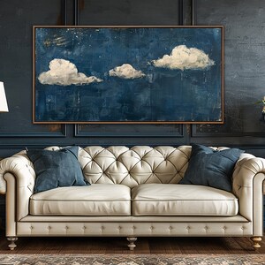 May include: A white tufted sofa with two blue pillows sits in front of a dark blue wall with a framed painting of white clouds on a blue background. Two gold end tables with lamps and a vase of white flowers are on either side of the sofa.