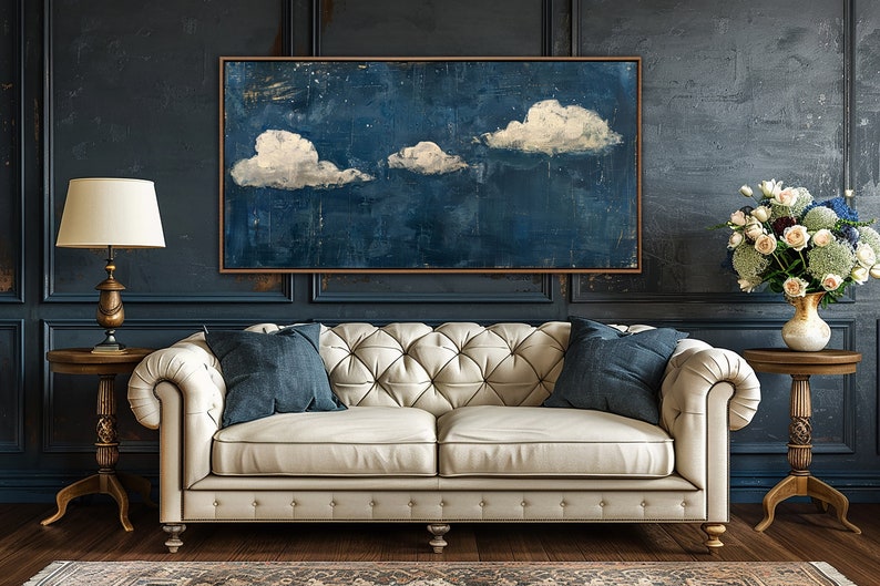 May include: A white tufted sofa with two blue pillows sits in front of a dark blue wall with a framed painting of white clouds on a blue background. Two gold end tables with lamps and a vase of white flowers are on either side of the sofa.