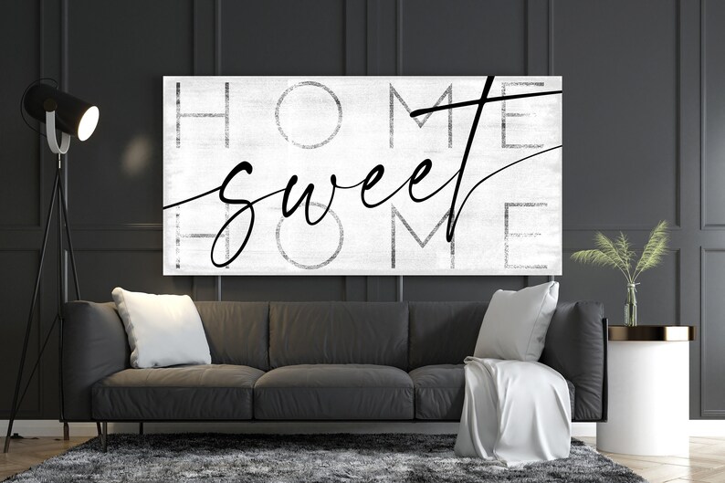 May include: A black and white canvas art piece with the words "HOME sweet HOME" in a mix of block and script fonts. The artwork hangs above a dark gray sofa with white pillows and a white throw blanket. A black floor lamp and side table complete the modern living room.