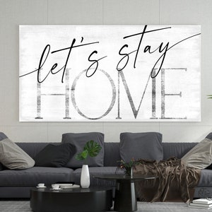 May include: A black and white canvas wall art print with the text "Let's stay home" in a modern calligraphy style. The print is displayed above a gray sofa in a living room setting.