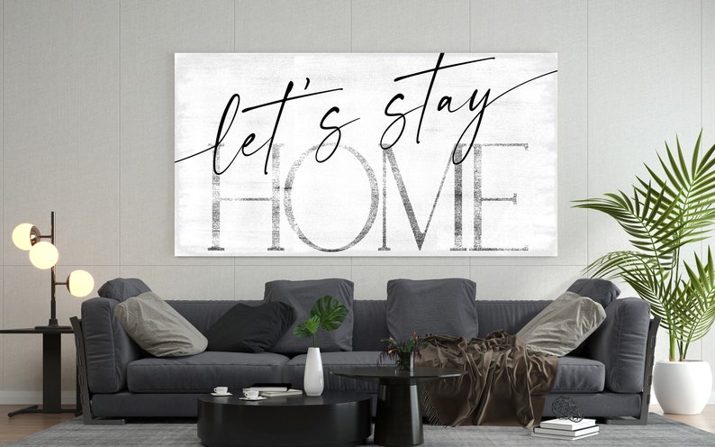 May include: A black and white canvas wall art print with the text "Let's stay home" in a modern calligraphy style. The print is displayed above a gray sofa in a living room setting.