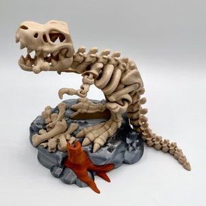 Puede incluir: Un modelo de esqueleto de dinosaurio impreso en 3D de color beige con una caja torácica y una cola detalladas. El dinosaurio está en posición de cuclillas sobre una base de roca gris con un tocón de árbol marrón.