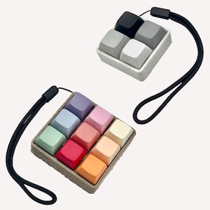 Può includere: Due fidget toy a forma di tasti quadrati con cinturini neri da polso. Uno presenta quattro tasti in nero, bianco e sfumature di grigio su una base bianca. L'altro presenta nove tasti in colori pastello e vivaci, tra cui viola, rosa, blu, verde, rosso, arancione e giallo su una base beige.