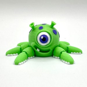 May include: A 3D-printed green monster figurine with one large eye, two small antennae, and multiple tentacle-like appendages. The eye is white with a blue iris and pupil. The monster has a smiling mouth and purple spots on its head.