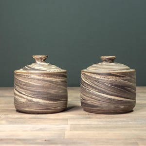 Op de afbeelding: Twee keramische potten met deksels, beide met een swirlend bruin en wit marmerpatroon. De potten staan op een houten oppervlak.