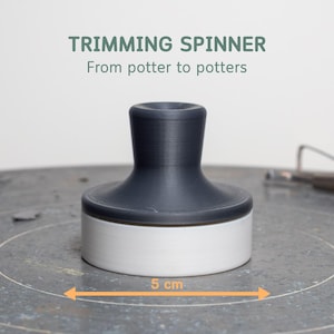 May include: A trimming spinner for pottery, with a dark gray top and a white base. The spinner is approximately 2 inches in diameter. The text "TRIMMING SPINNER" and "From potter to potters" is displayed above the spinner.