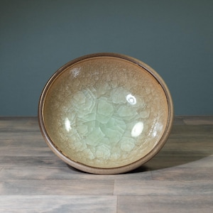 Könnte beinhalten: Eine runde Keramikschale mit Craquelé-Glasur. Die Schale hat eine hellgrüne Mitte, die in einen beigen Rand übergeht. Die Schale steht auf einer Holzoberfläche.