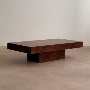 May include: A rectangular coffee table with a dark brown, burl wood finish. The table has a low profile, with a thick top and a central, rectangular base. The wood grain is visible, adding texture to the piece. The table is approximately 60 inches long and 30 inches wide.