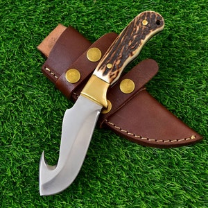 May include: A hunting knife with a curved blade and a handle made of bone-like material and dark brown accents. The knife has a brass bolster and is housed in a brown leather sheath with brass rivets.