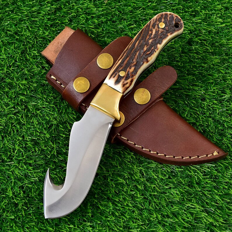 May include: A hunting knife with a curved blade and a handle made of bone-like material and dark brown accents. The knife has a brass bolster and is housed in a brown leather sheath with brass rivets.
