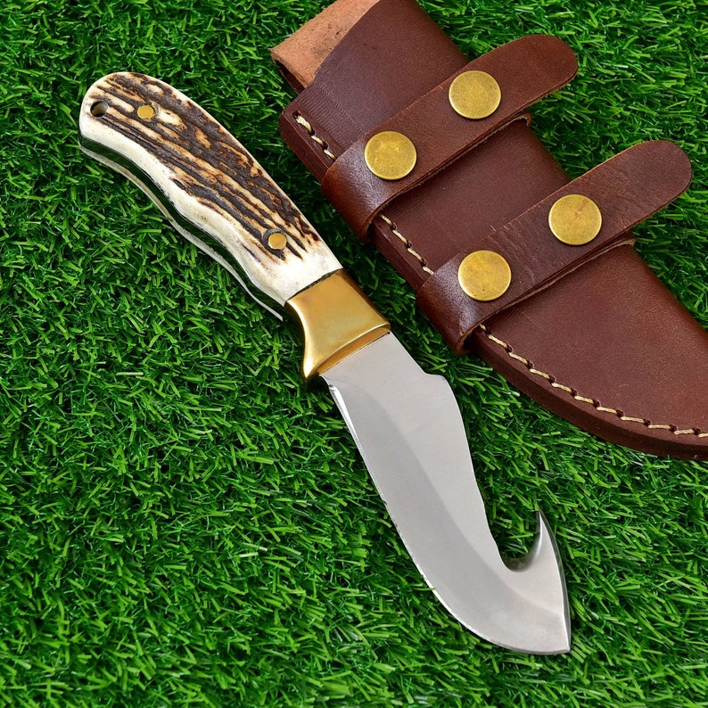 May include: A hunting knife with a curved blade and a bone handle. The knife has a brass bolster and is paired with a brown leather sheath with brass rivets. The knife is set against a green background.