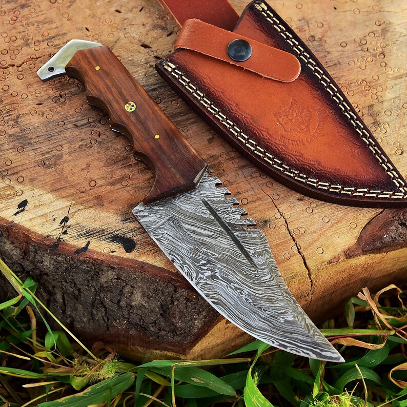 May include: A Damascus steel hunting knife with a wooden handle and a leather sheath. The knife has a patterned blade and a serrated edge.