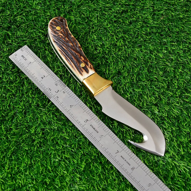 May include: A hunting knife with a stainless steel blade and a hook at the tip. The handle features a brown and cream pattern with brass accents. A metal ruler is placed next to the knife.