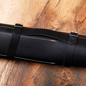 May include: A black leather knife roll with a handle and two straps with buckles. The roll is rolled up and sitting on a wooden surface.