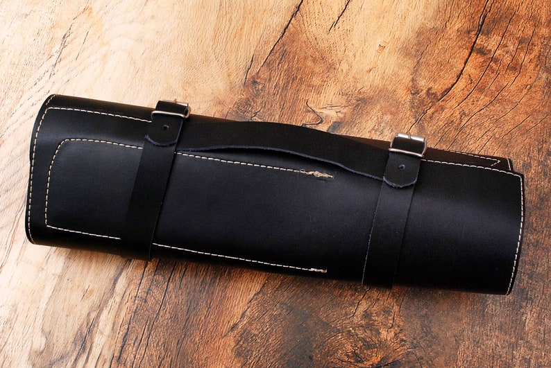 May include: A black leather knife roll with a handle and two straps with buckles. The roll is rolled up and sitting on a wooden surface.