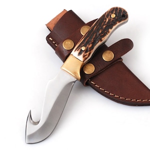 May include: A hunting knife with a curved blade and a brown and white antler handle. The knife is in a brown leather sheath with brass rivets. The blade is stainless steel.