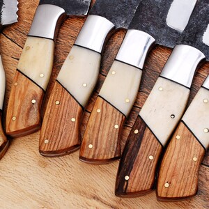 May include: A set of eight knives with wooden handles and white bone accents. The knives have a rustic, handmade look and are arranged in a row on a wooden surface.