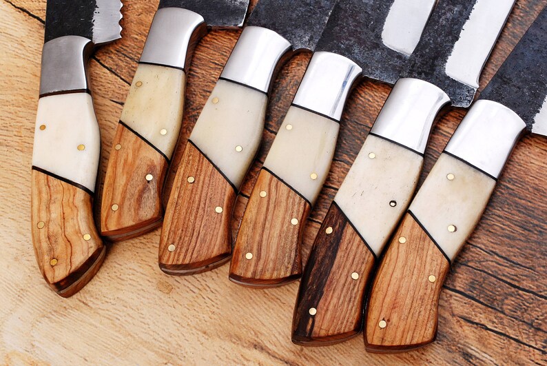 May include: A set of eight knives with wooden handles and white bone accents. The knives have a rustic, handmade look and are arranged in a row on a wooden surface.