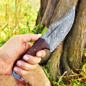 May include: A close-up of a hand holding a hunting knife with a wooden handle and a patterned steel blade.