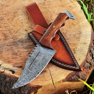 May include: A Damascus steel hunting knife with a wooden handle and a leather sheath. The knife has a serrated edge and a decorative pattern on the blade.