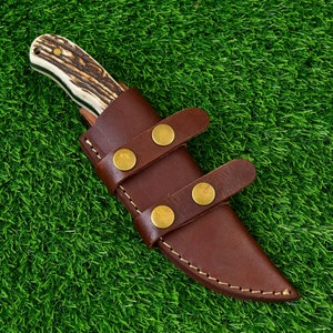 May include: A hunting knife with a textured antler handle and a brown leather sheath. The sheath has two straps secured with brass-colored rivets. The knife is designed for outdoor use and survival.