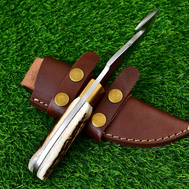 May include: A hunting knife with a curved blade and a handle made of bone-like material. The knife is in a brown leather sheath with brass rivets. The knife is designed for outdoor use.