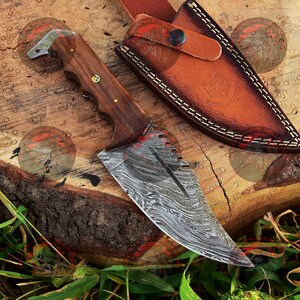 May include: A Damascus steel knife with a wooden handle and a leather sheath. The knife has a unique pattern on the blade and a serrated edge.
