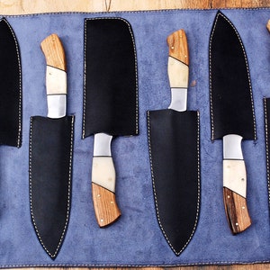 May include: A set of six knives with wooden handles and white bone accents are displayed in a blue leather roll. The knives are arranged in a row, with the handles facing up. The roll has a stitched border and is made of a soft, supple leather.