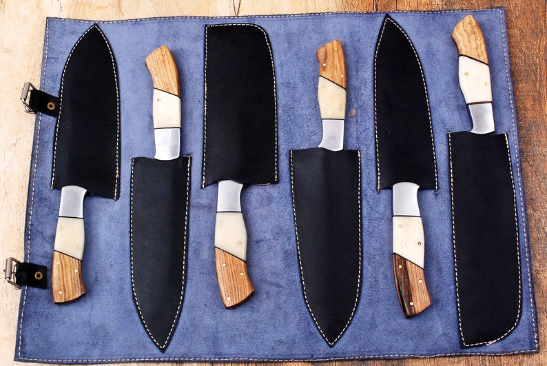 May include: A set of six knives with wooden handles and white bone accents are displayed in a blue leather roll. The knives are arranged in a row, with the handles facing up. The roll has a stitched border and is made of a soft, supple leather.