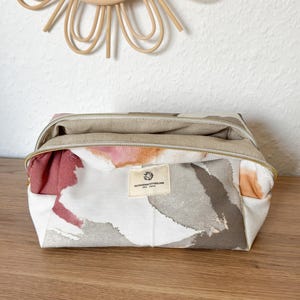 Op de afbeelding: Een rechthoekige make-uptas met een bloemenpatroon in tinten wit, grijs, roze en oranje. De tas heeft een gouden rits en een klein label met tekst. De tas staat op een houten oppervlak.
