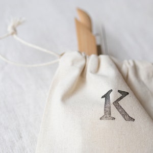May include: A white drawstring bag with a black letter "K" printed on the front. The bag is made of natural fabric and has a simple design.