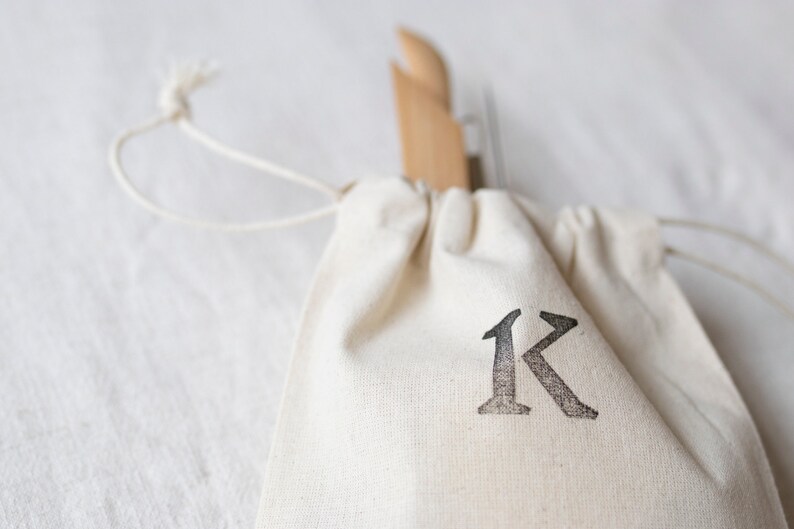 May include: A white drawstring bag with a black letter "K" printed on the front. The bag is made of natural fabric and has a simple design.