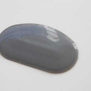 Könnte beinhalten: Eine glatte, ovale Pfütze aus dunkelgrauer Farbe vor einem weißen Hintergrund. Die Farbe hat einen glänzenden Glanz, der das Licht reflektiert und Glanzlichter erzeugt. Das Bild ist eine Nahaufnahme, die Textur und Farbe hervorhebt.