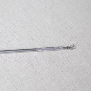 Puede incluir: Una herramienta plateada con un mango texturizado y un grupo de alambres finos en un extremo. La herramienta está diseñada para un propósito específico, probablemente relacionado con la limpieza o los detalles. El fondo es una tela lisa de color blanquecino.