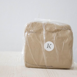 Può includere: Un blocco di argilla beige avvolto in plastica trasparente con un'etichetta circolare bianca su cui è scritto "K".
