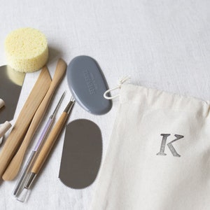 May include: A set of pottery tools in a white canvas bag with a black letter K. The tools include a wooden paddle, a metal scraper, a metal loop tool, a sponge, and a gray silicone tool.
