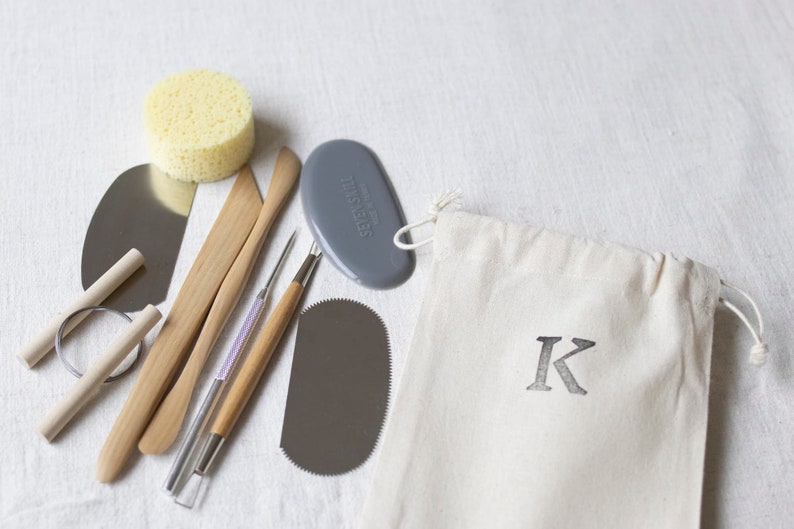 May include: A set of pottery tools in a white canvas bag with a black letter K. The tools include a wooden paddle, a metal scraper, a metal loop tool, a sponge, and a gray silicone tool.